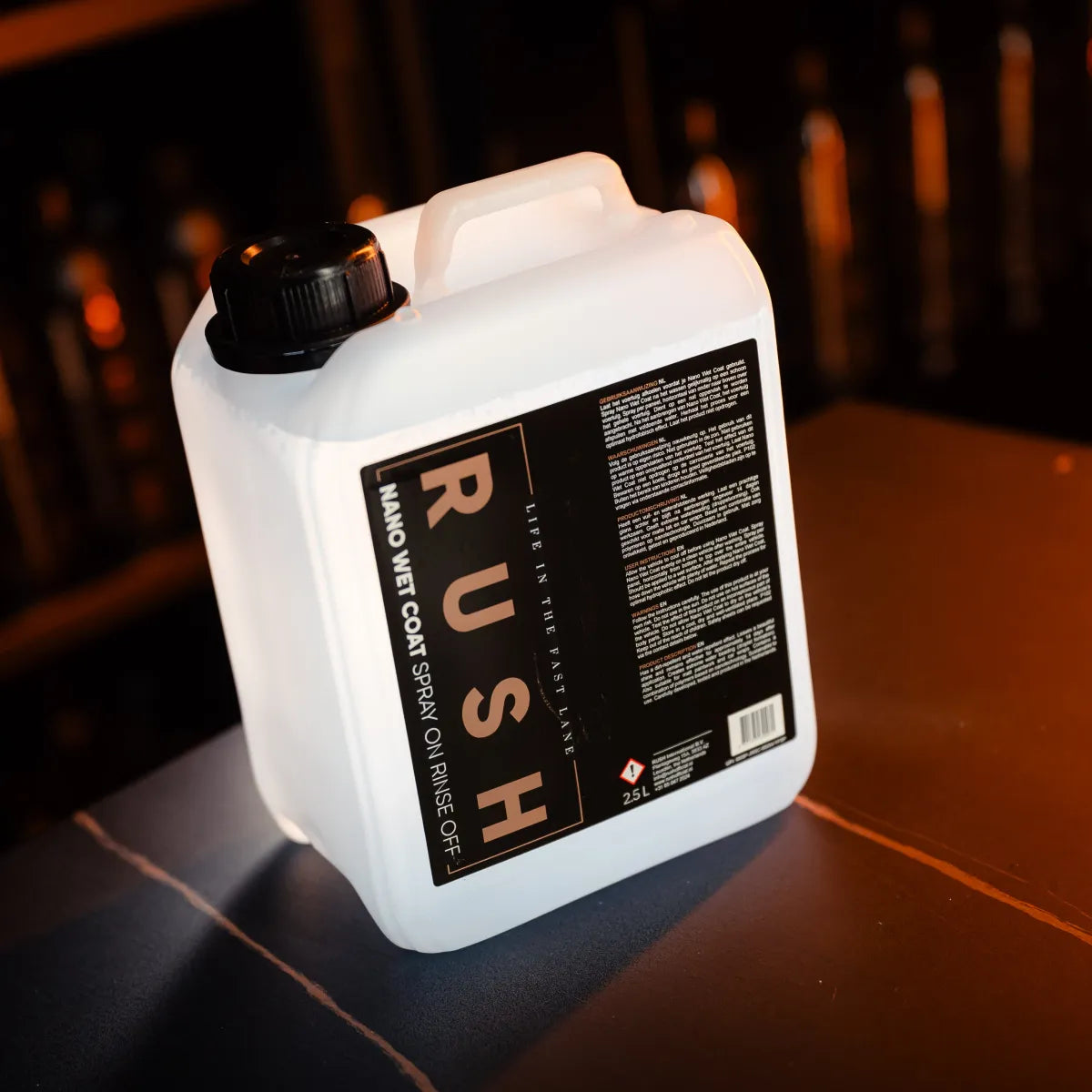A white 2.5-liter plastic container of Nano Wet Coat 2,5 liter rests on an orange surface, with a black label showing details. The dim, out-of-focus background contrasts with the product in the foreground.