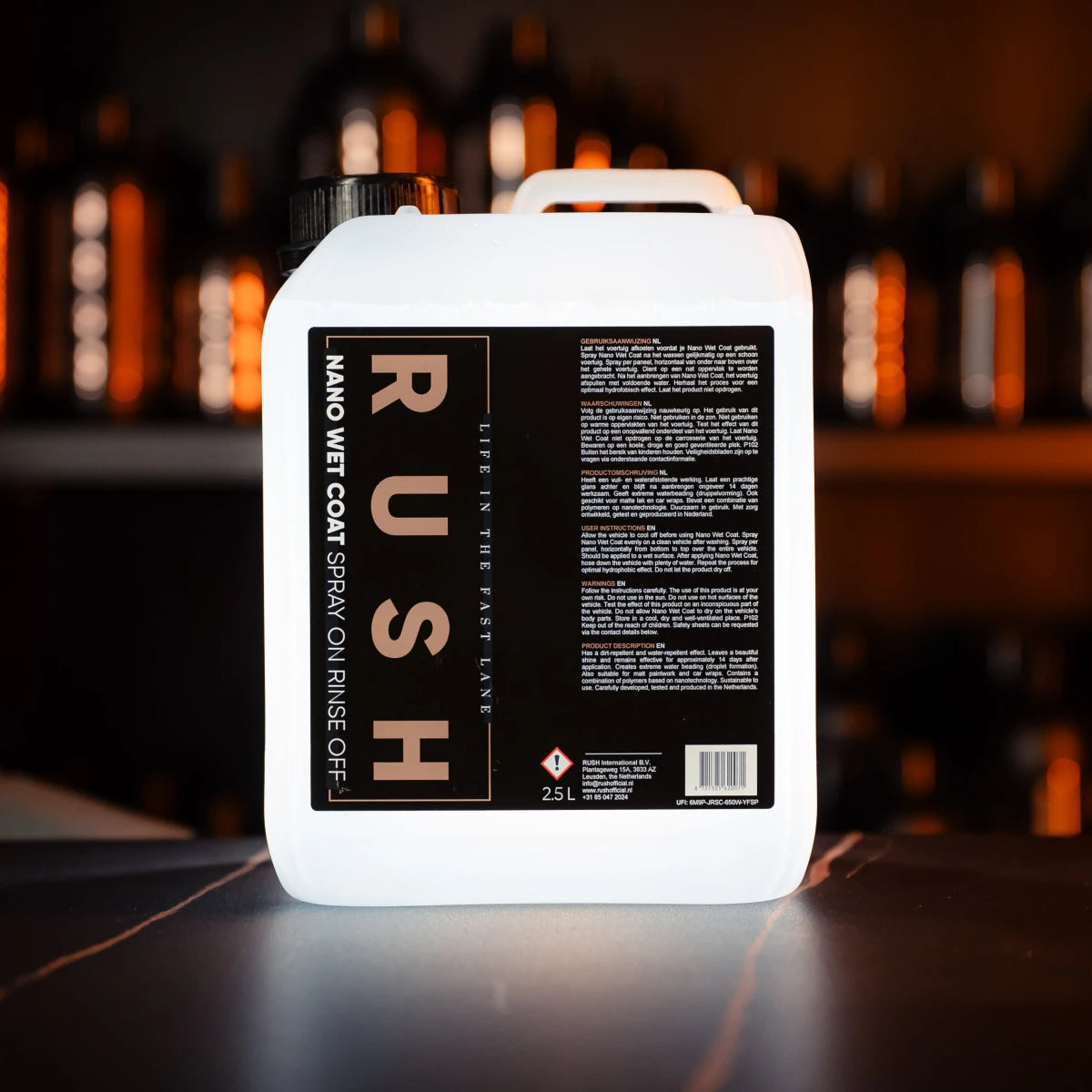 A white 2.5-liter container labeled Nano Wet Coat 2,5 liter sits on a counter with text and instructions on its black label, against a softly lit background featuring blurred bottles.
