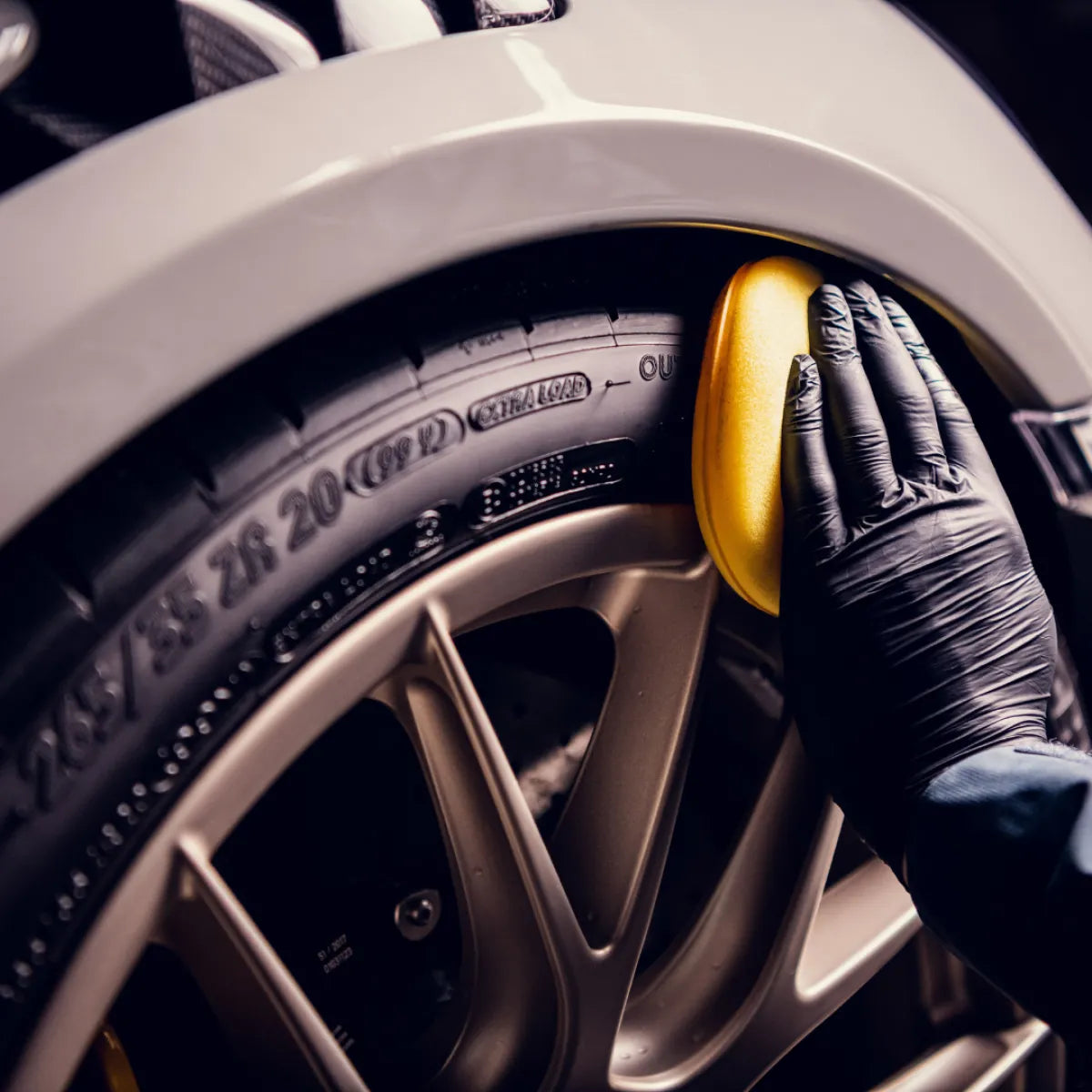 Een gehandschoende hand brengt bandenglans aan op een autoband met een gele spons, gericht op de band en het lichtmetalen wiel.