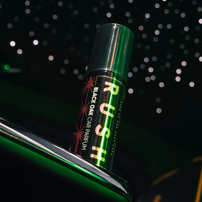 Een blikje met het label RUSH BLACK OAK AUTOPARFUM rust op het interieur van een auto en verspreidt een langdurig ruikende geur te midden van wazige lichten.