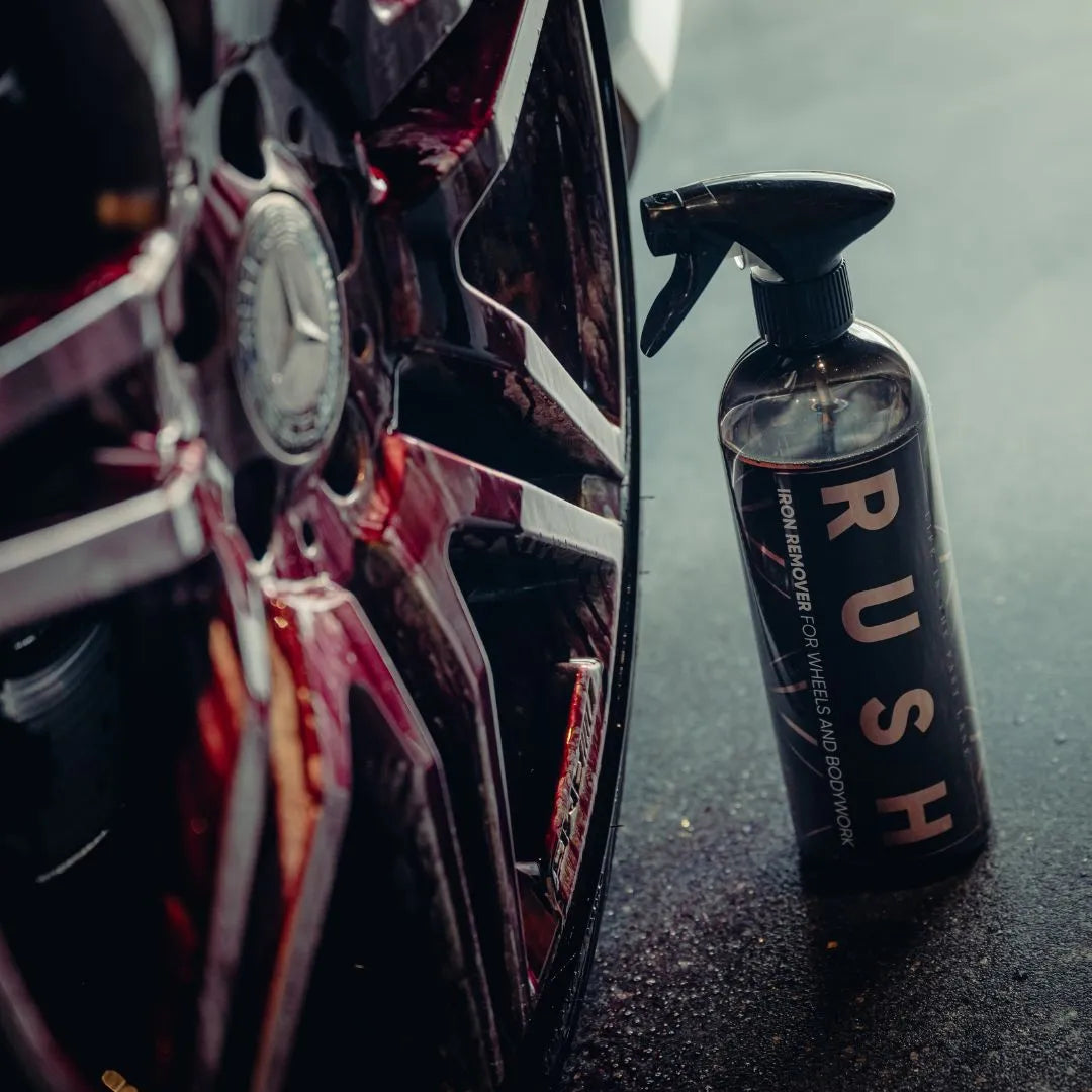 Een fles RUSH IJzerverwijderaar velgenreiniger staat bij een autowiel met embleem op een donker, reflecterend oppervlak.
