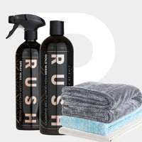 Twee zwarte flessen met het label RUSH, perfect voor je spraywaxroutine, staan naast gevouwen handdoeken op een lichtgrijze achtergrond.