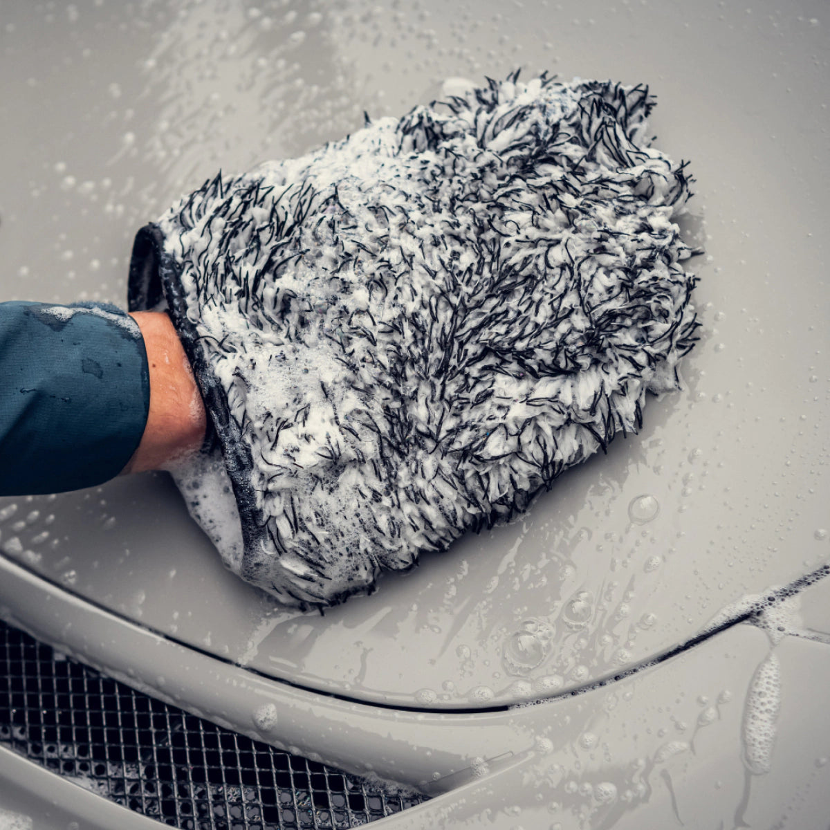 Een persoon gebruikt een microvezel washandschoen met groot absorberend vermogen om de motorkap van een lichtgekleurde auto te wassen.