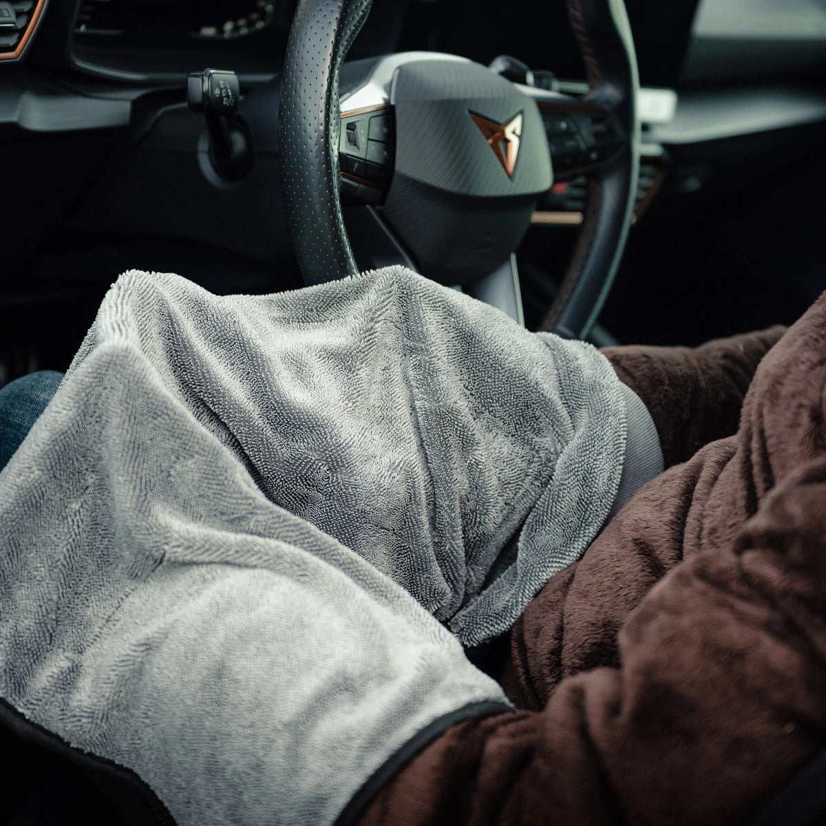 Een persoon zit in een auto met een grijze deken en bruine jas, klaar om de RUSH Window Clear Winter Kit te gebruiken om de voorruit ijsvrij te maken.