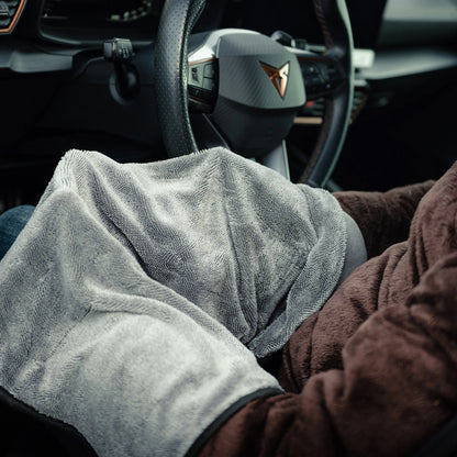 Een persoon zit in een auto met een grijze deken en bruine jas, klaar om de RUSH Window Clear Winter Kit te gebruiken om de voorruit ijsvrij te maken.