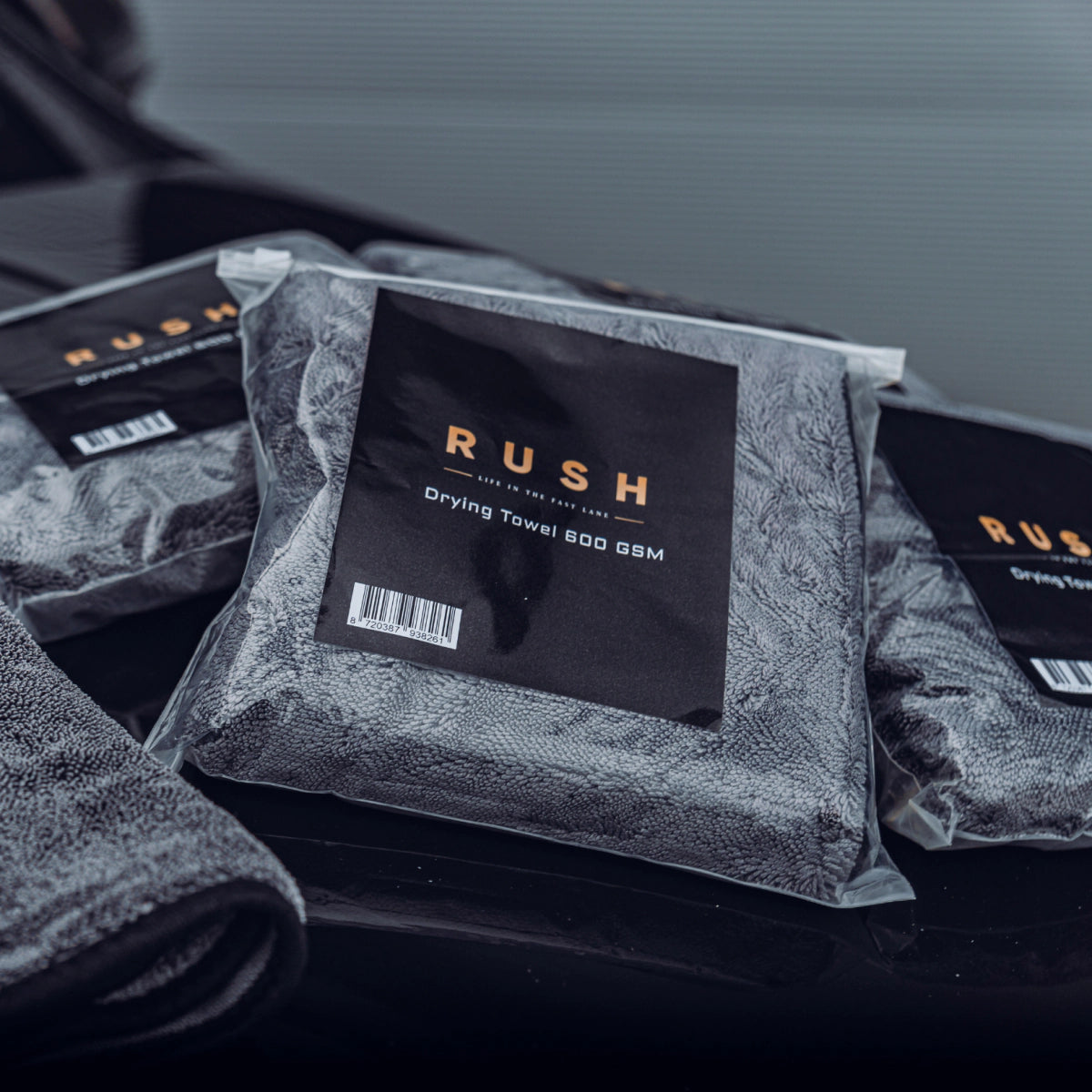 Drie verpakte grijze droogdoeken 600 GSM met het label RUSH worden weergegeven op een donker, glanzend oppervlak - perfect als droogdoek auto.