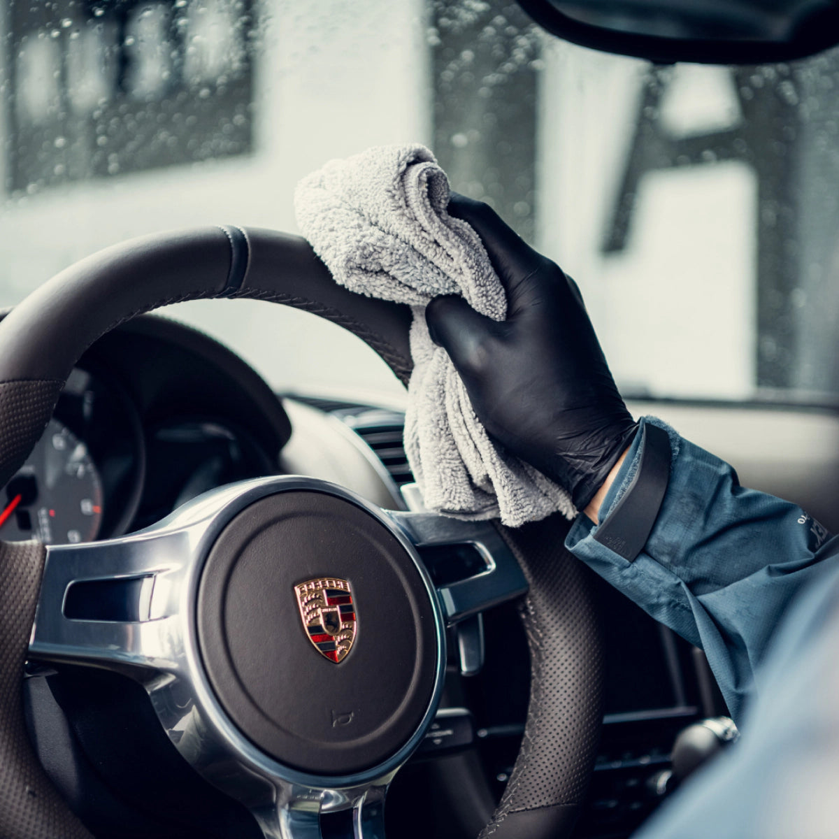 Een persoon met een zwarte handschoen gebruikt interieurspray om het stuur van een Porsche af te vegen met een grijze doek in de auto op een regenachtige dag.