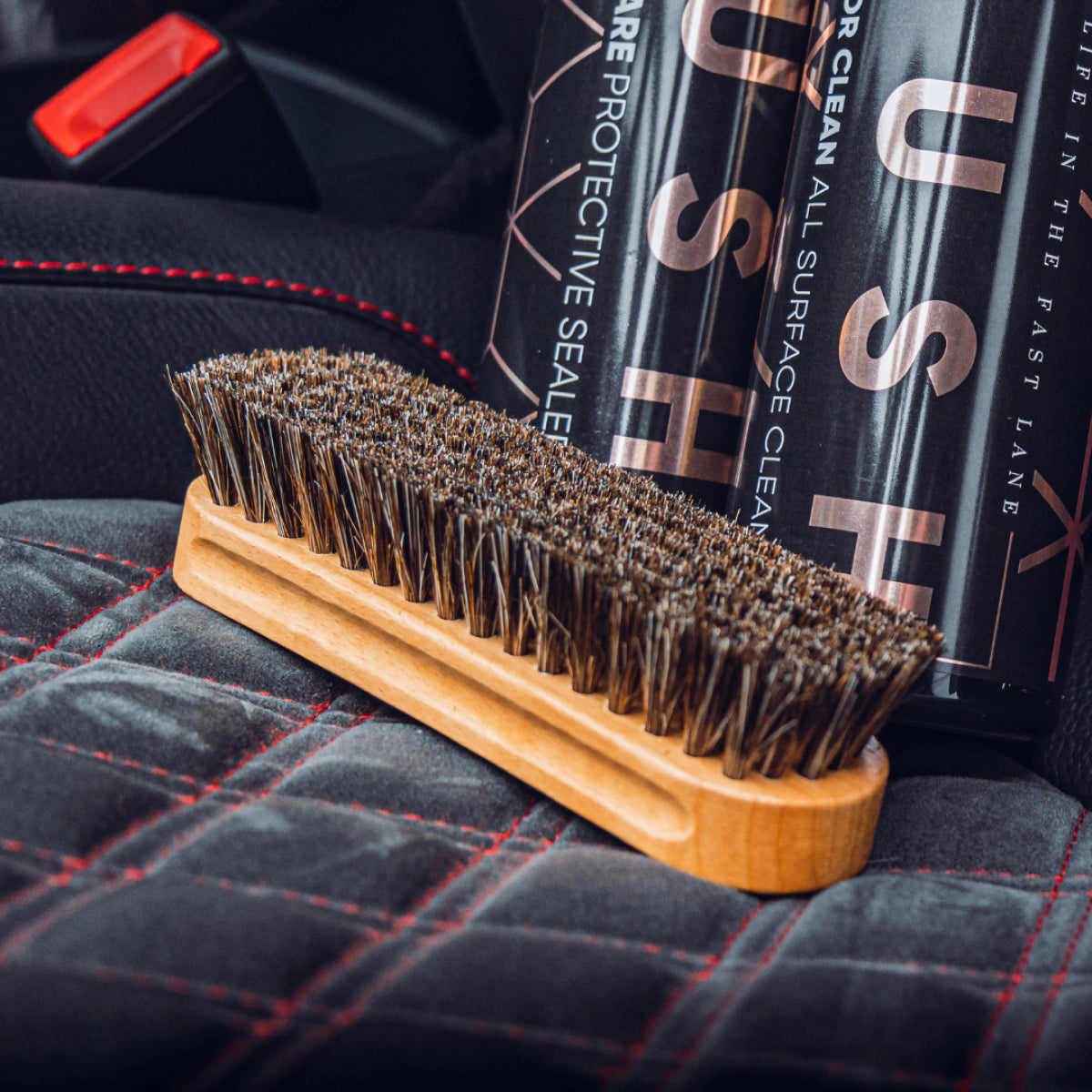 A close-up of the Leather Cleaning Brush with dark bristles rests on quilted black car seat upholstery, showing it as the ideal tool for cleaning leather, with cleaning product bottles visible in the background.
