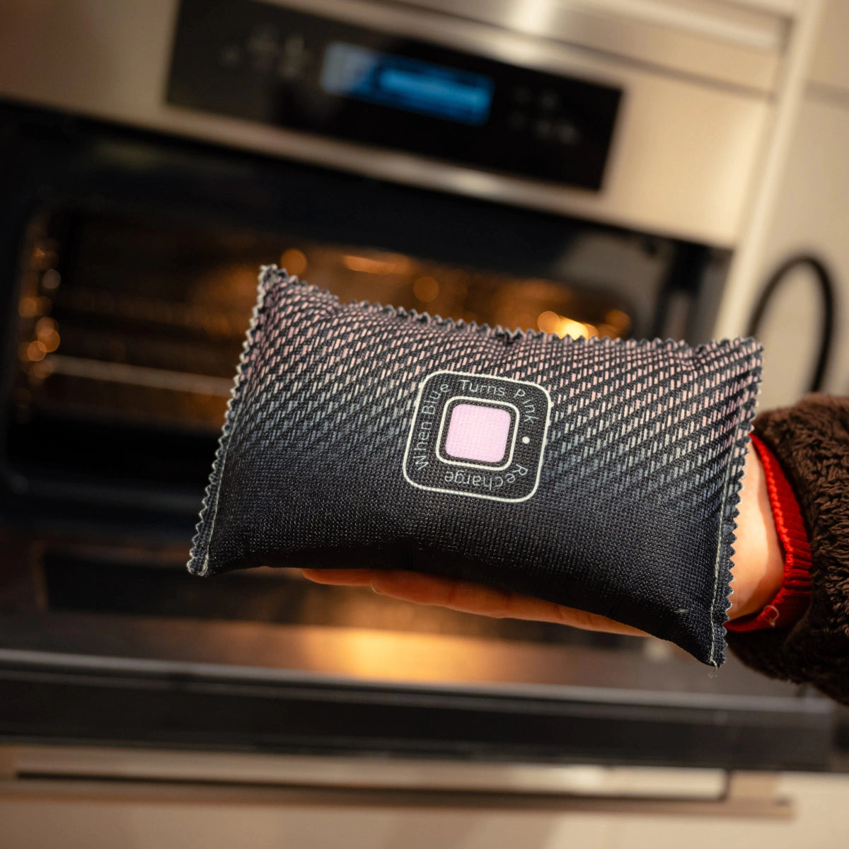 A hand holds a rectangular fabric pouch labeled "Turns" and "Change" in front of an open oven. This woven-texture auto dehumidifier keeps interiors dry—a true Winter Essentials Package for your car.