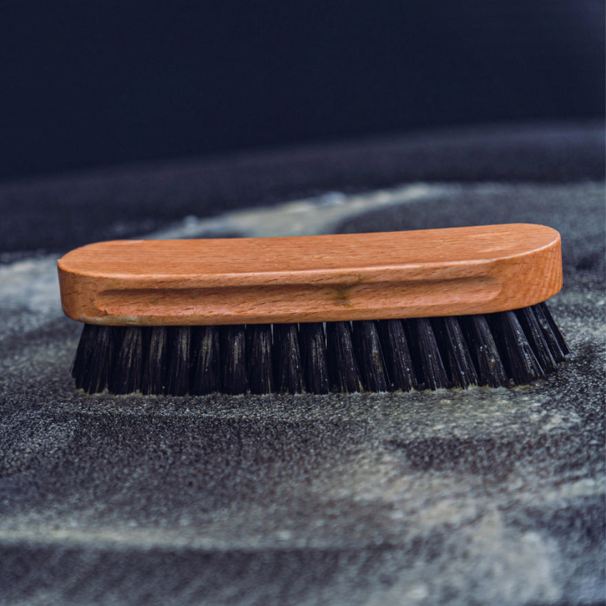 Een textielborstel met zwarte nylon borstelharen rust op donkere stof, die tekenen vertoont van recent auto interieur reinigen.