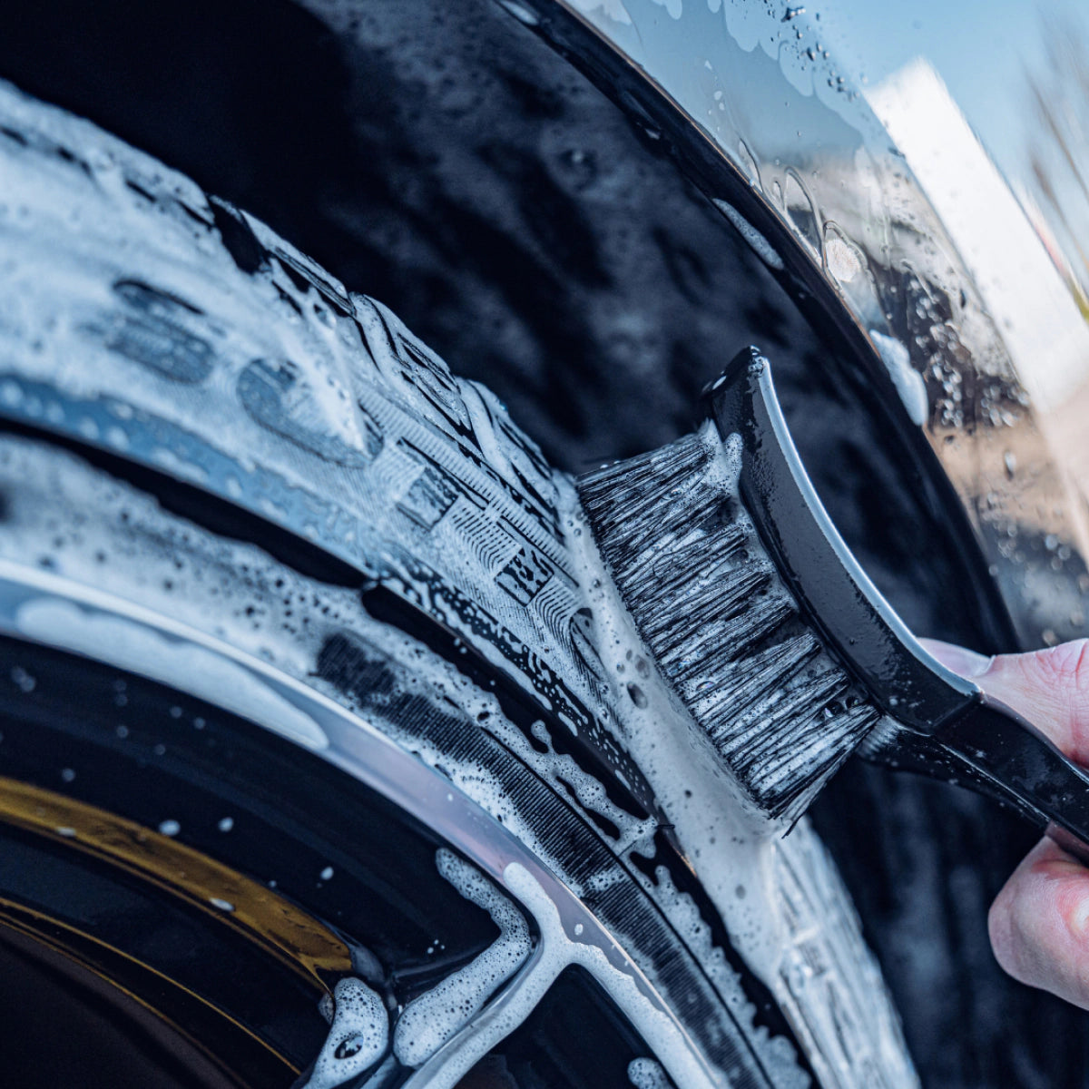 A close-up of a hand scrubbing a car tire with the Tire Brush, creating soap suds on the black rubber surface—demonstrating effective tire cleaning.