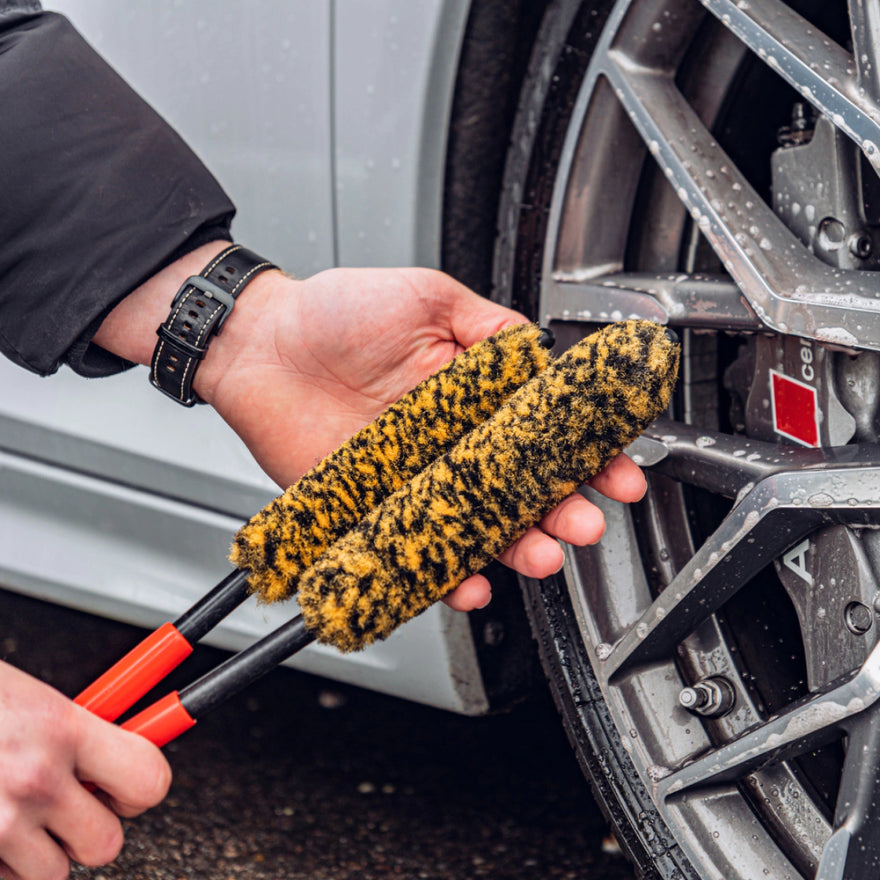 Een persoon houdt een Wheel Woolie Set bij het lichtmetalen wiel van een zilverkleurige auto, terwijl hij bezig is met velgen reinigen.