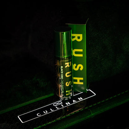 Een flesje en doosje Rush Black Oak Cardamom auto parfum staan uitgestald op een oppervlak met het label Cullinan, verlicht met groen licht.