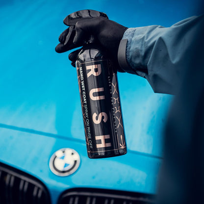 Een gehandschoende hand houdt een fles met het opschrift RUSH spray wax voor de motorkap van een blauwe BMW en belooft extreme waterbeading bij elk gebruik.