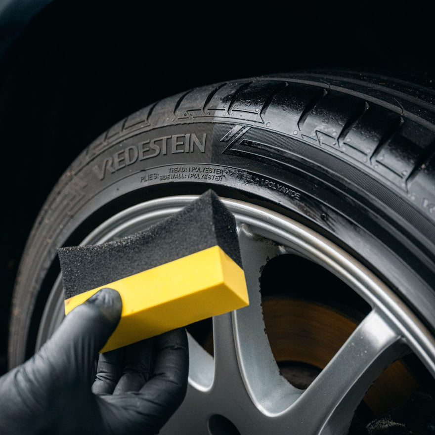 Een gehandschoende hand houdt een geel met zwarte spons vast bij een schone Vredestein autoband, perfect voor bandenglans en rubber verzorging.