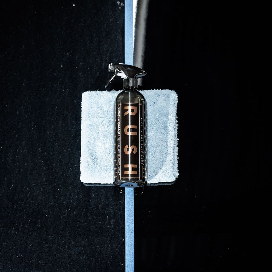 Een spuitfles met het label RUSH, perfect voor de winterbescherming van autos, rust op een blauwe microvezeldoek op een zwart oppervlak met een lichtblauwe streep.
