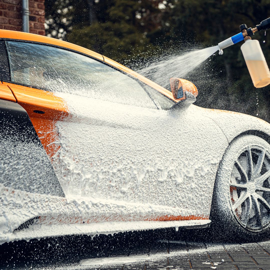 Iemand spuit sneeuwschuim op een oranje sportwagen met een hogedrukreiniger tijdens een wasbeurt.