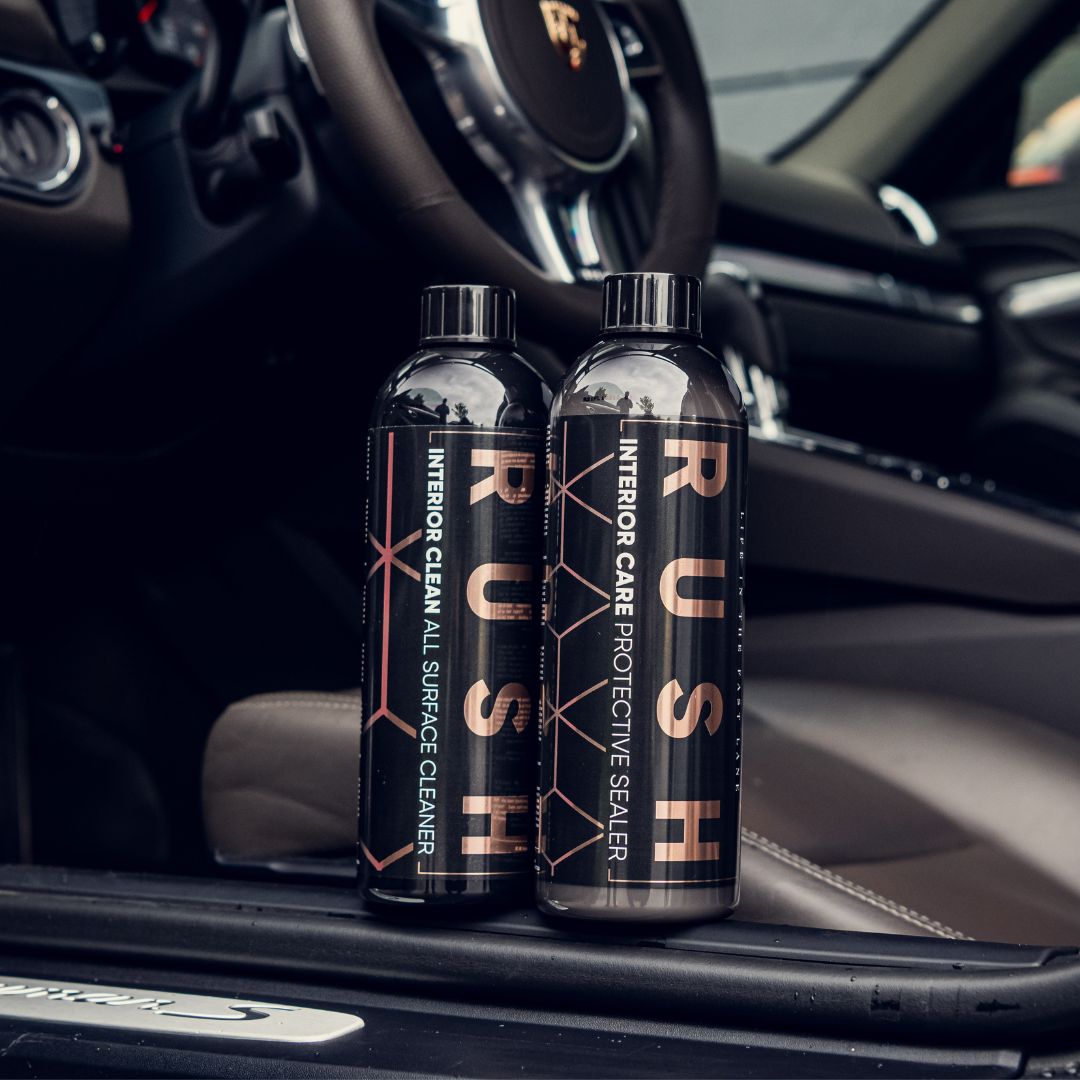 Two bottles from the Detailing Package PLUS | 23-delig rest on the door frame, showcasing the car’s beige seats and steering wheel. The labels read All Surface Cleaner and Protective Sealer.