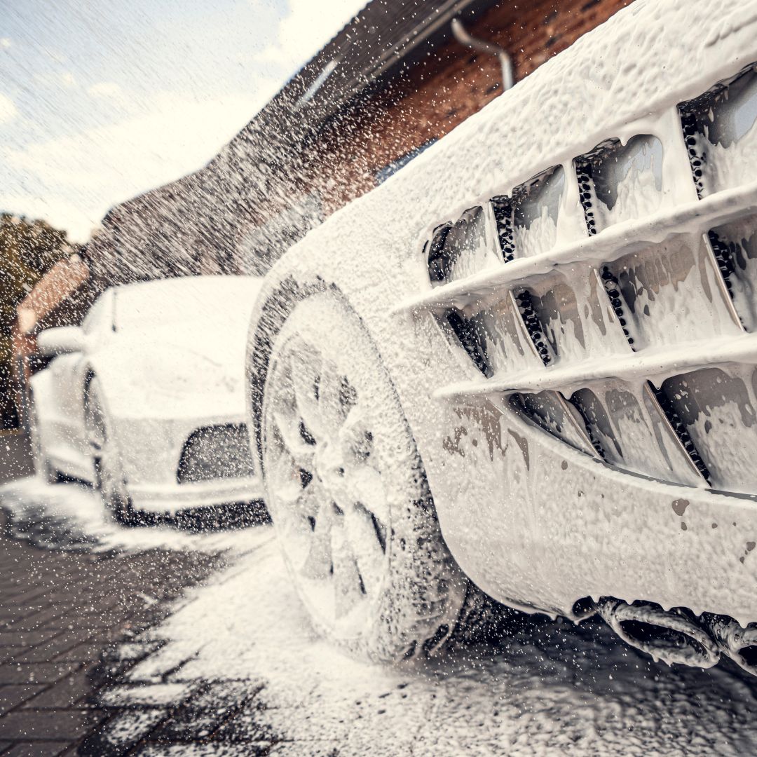 Close-up van twee sportautos bedekt met een dikke schuimlaag van sneeuwschuim tijdens een autowasbeurt, met spuitend sop op de voorgrond.