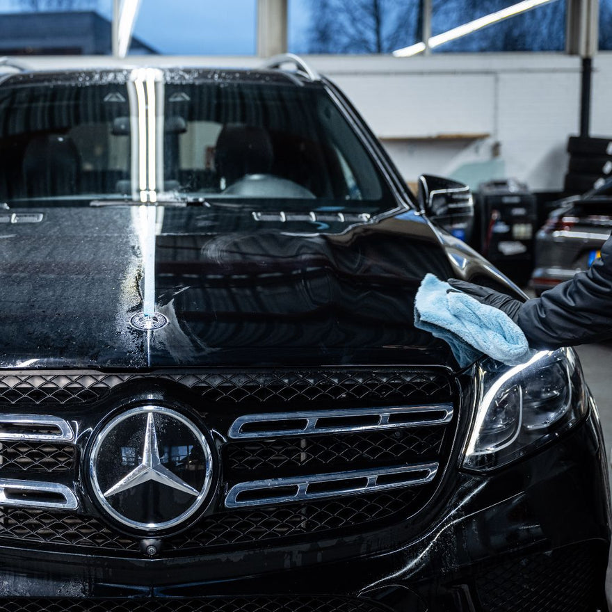A person uses a blue microfiber cloth to wipe the hood of a shiny black Mercedes-Benz SUV in a garage, applying the Ceramic Sealant Package for extra paint protection.