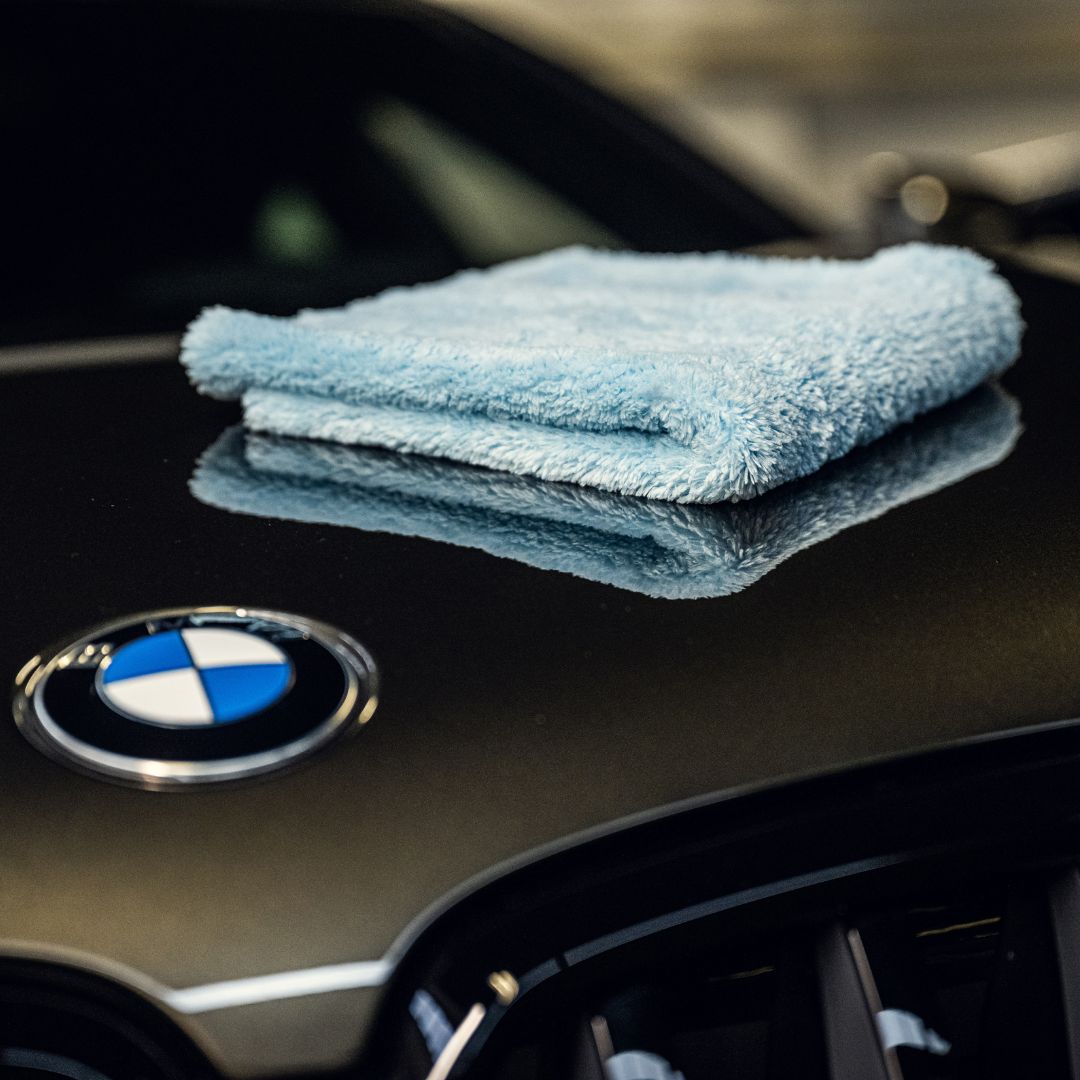 A folded light blue microfiber towel sits on the shiny black hood of a BMW, next to its emblem—highlighting the stunning gloss and lak bescherming you can achieve with 1+1 GRATIS Spray Wax.