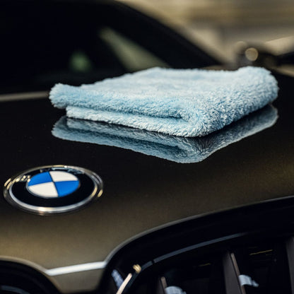 A folded light blue microfiber towel sits on the shiny black hood of a BMW, next to its emblem—highlighting the stunning gloss and lak bescherming you can achieve with 1+1 GRATIS Spray Wax.