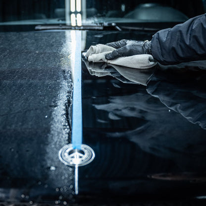 A gloved hand wipes a cloth over a black car hood, cleaning and polishing the surface. The Ceramic Sealant Package boosts shine, as light reflects off the finish to highlight the difference between protected and unprotected areas.