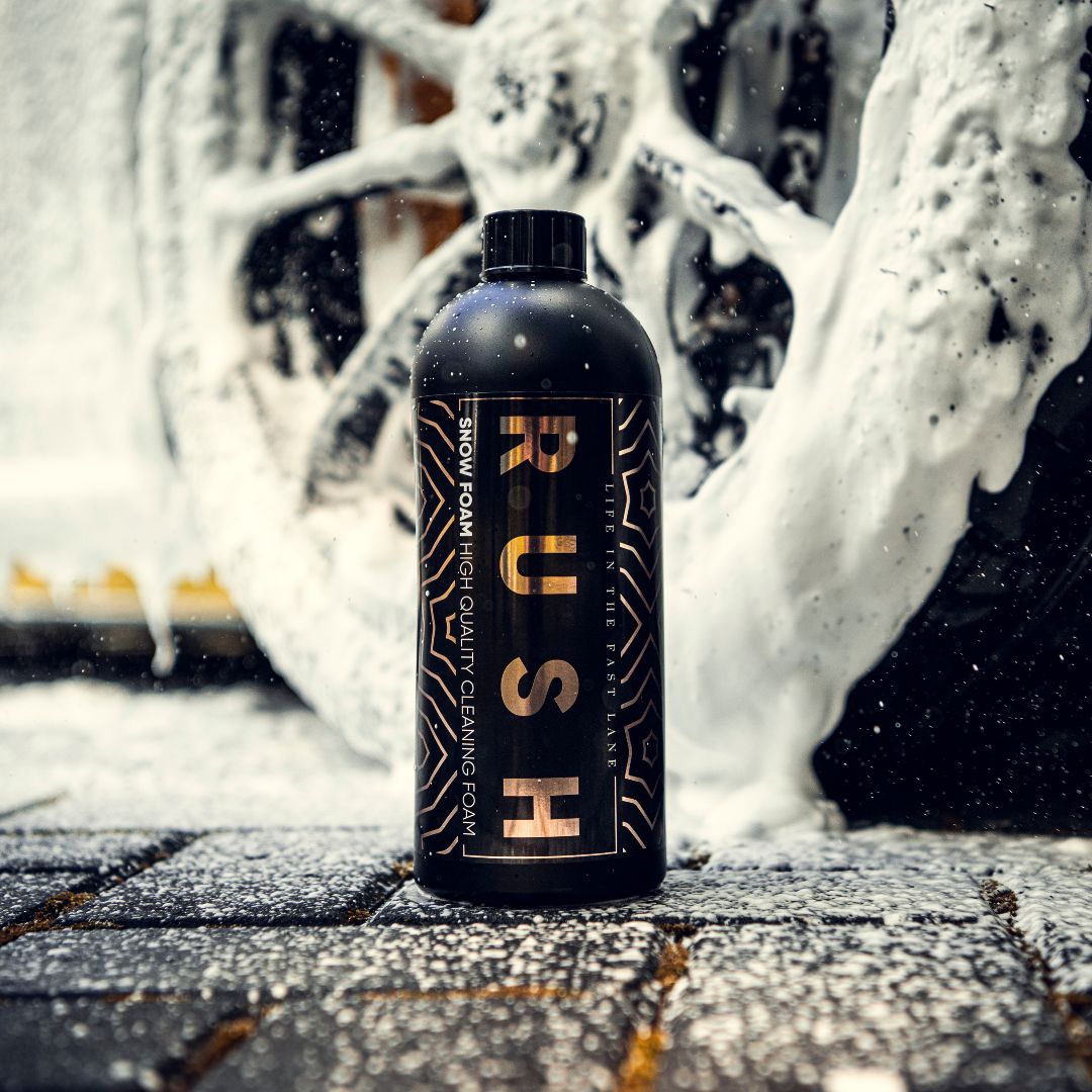 Een zwarte fles met het label RUSH Snow Foam staat op een nat wegdek, omringd door een dikke schuimlaag en een wiel met schuim op de achtergrond.