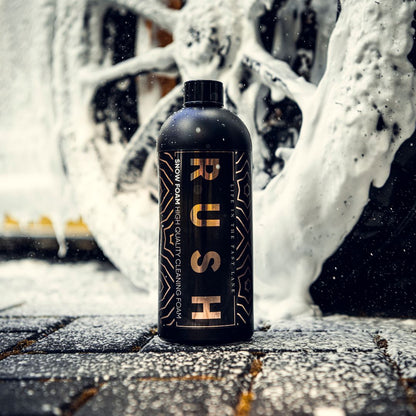 Een zwarte fles met het label RUSH Snow Foam staat op een nat wegdek, omringd door een dikke schuimlaag en een wiel met schuim op de achtergrond.