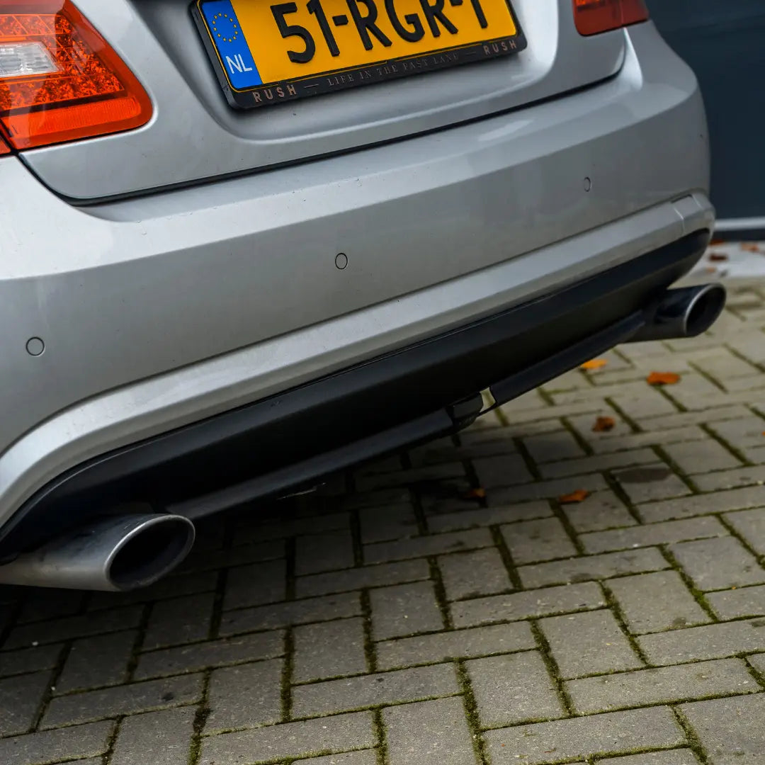 Een close-up van de achterbumper en dubbele uitlaatpijpen van een zilverkleurige auto accentueert kunststof exterieur herstellen op klinkerbestrating met een Nederlands bord.