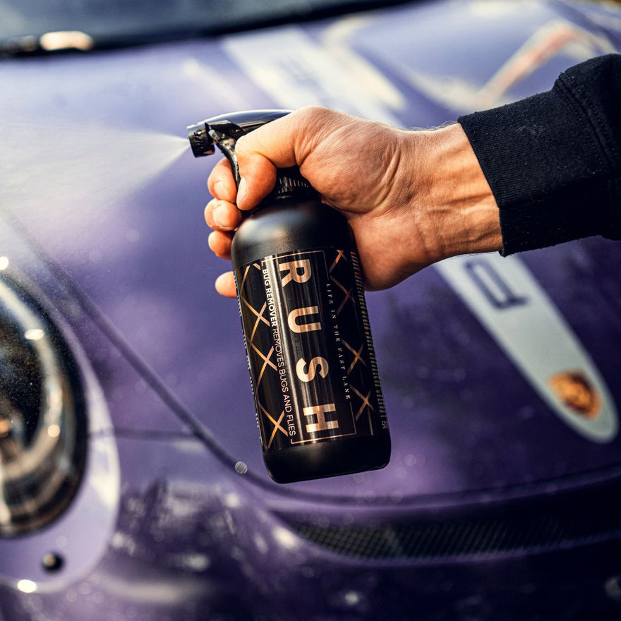 Een hand houdt een fles insectenverwijderaar met de naam Rush voor de motorkap en koplamp van een paarse sportwagen.