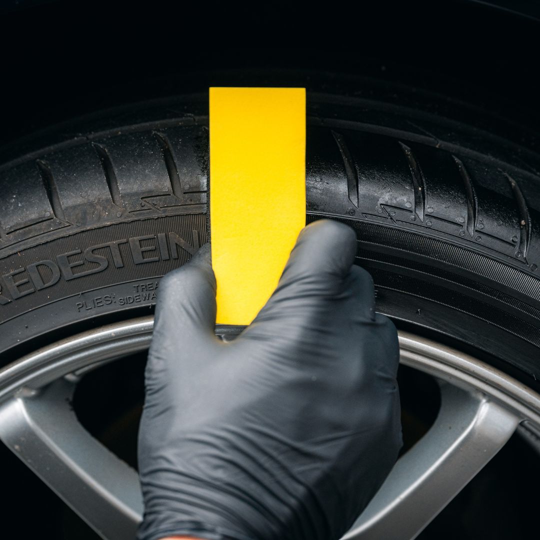 Een gehandschoende hand houdt een gele kaart bij een autoband om de profieldiepte te controleren als onderdeel van de rubberen verzorging of bandenglansroutine.