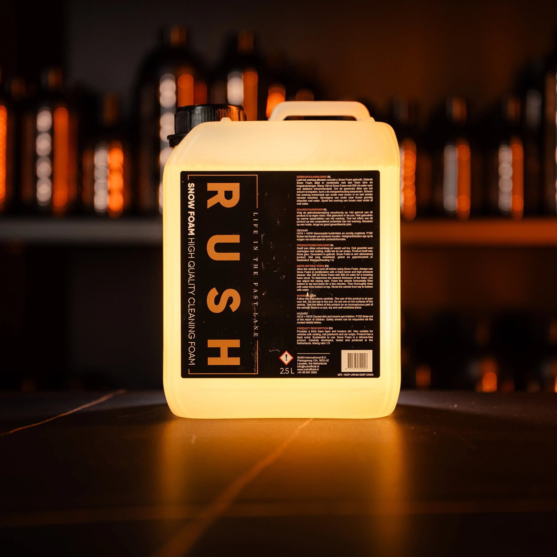 Een 25-liter container met het label RUSH Snow Foam High Quality Cleaning Foam wordt verlicht op een donkere plank met flessen op de achtergrond.
