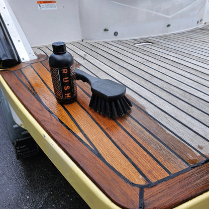 Een fles met het label RUSH en een schoonmaakborstel rusten op een houten bootdek, klaar om teakhout te reinigen en teakhout kleur te herstellen.