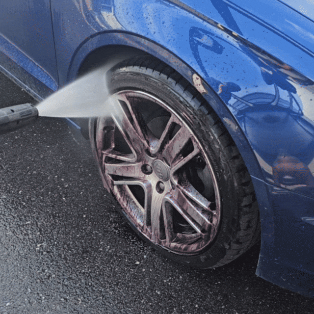 A close-up of a blue car's wheel being cleaned with a high-pressure water jet and the Black Friday Package, washing off purple brake dust or cleaner from the alloy rim on wet black asphalt.